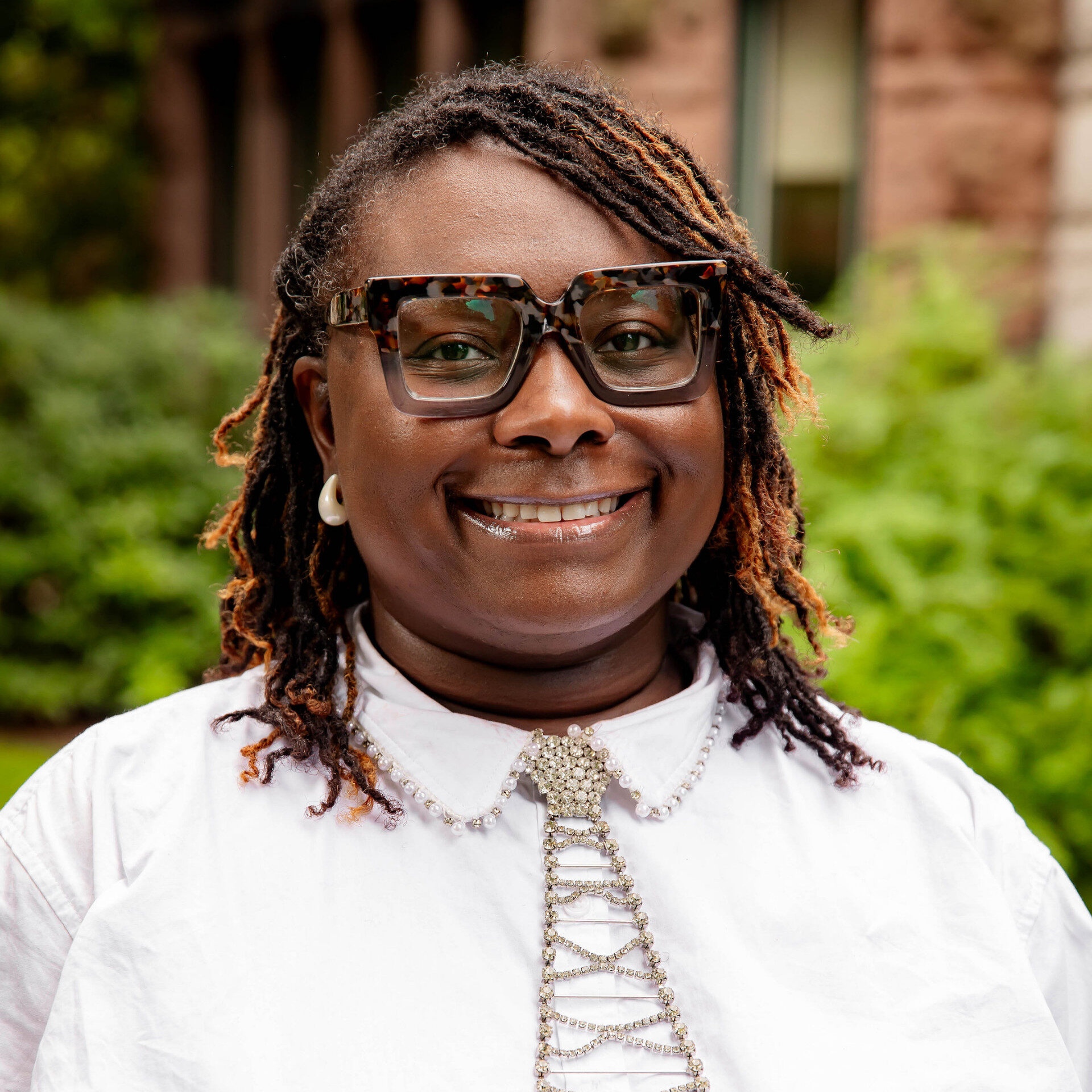 Smiling woman with locs, tortoiseshell glasses, and rhinestone tie